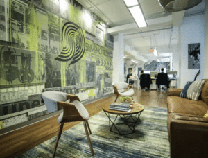 Modern office lounge area with contemporary chairs, a coffee table with books, a leather sofa, and a mural wall; employees work at desks in the background, creating a symphonic blend of collaboration and comfort.